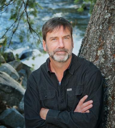 Poet Derek Sheffield, arms crossed, stands next to a tree with a river in the background.