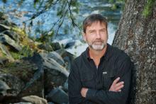 Poet Derek Sheffield, arms crossed, stands next to a tree with a river in the background.