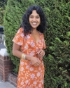 Woman with curly black hair in an orange dress standing in front of green hedge