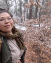 woman with brown hair in a top knot stands to the left-hand side of the photo frame, dressed in hiking gear. on the right hand side of the photo is a nature scene with sparse trees and snow on the ground