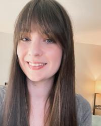 Kait is a white woman with long brown hair and bangs, wearing a grey shirt and smiling against a plain background.