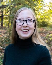 A blonde, white woman with glasses smiling at the camera in a forest setting