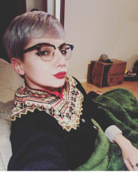 a person with bleached, short hair stares sideways at the camera. They are wearing a colorful sweater, red lipstick, cat's eyeliner, and black and gold framed glasses 