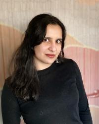 A photo of nanya in a black shirt standing in front of a pink, orange, and beige background. 