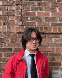 Person seen from the chest up, wearing a white shirt, black tie, and a red jacket against a background of bricks.