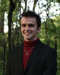 Dark-haired Caucasian man wearing a red turtleneck and black blazer smiles in front of a forest background.