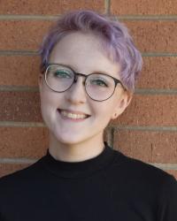 A head and shoulders portrait of a white person with short purple hair and round glasses. They are wearing a black turtleneck, and stand in front of a brick wall. They are smiling widely.