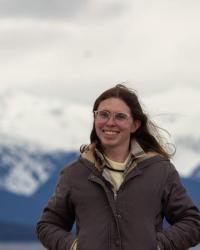 Olive, a white, transgender woman, looks a the camera. She wears a gray jacket, has long brown hair, and stands in front of a mountain range.