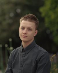 Medium length portrait with subject looking at the camera. A green foliage background is thrown out of focus by the shallow depth of field. Subject is wearing a button up oxford shirt and has short hair.