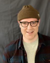 Man wearing a brown beanie, dark-framed glasses, and open flannel shirt over a light grey t-shirt stands smiling in front of a dark grey backdrop