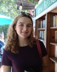 Madeleine Hearn, a white woman with curly brown hair, stands next to an outdoor bookshelf. 