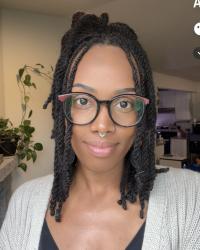 Female presenting person, brown skin, black glasses, plants in the background. 