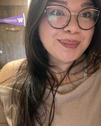 woman with brown hair and glasses sits at a desk, a university of washington flag is in the background