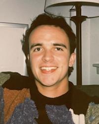 A young, clean-shaven white man with medium-length dark hair and a multicolored sweater grins at the camera.