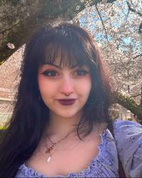  A selfie of a white woman with dark hair standing in front of cherry blossoms. She has a blue shirt on and dark purple lipstick. 