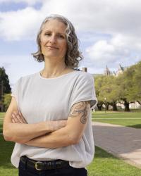 Headshot of brown and gray-haired woman crossing arms with green trees and grass in background