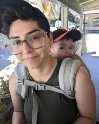 Woman with short hair and glasses smiling at the camera while wearing a toddler on her back.