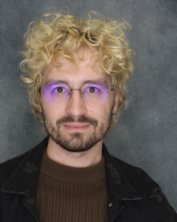 Face photo from the shoulders up of Alex Henry. They are bleach-blond haired, curly, with glasses on.