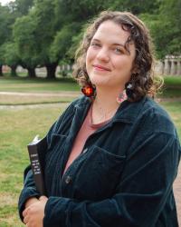 photograph of woman with curly hair in green jacket standing outside, holding a book and smiling