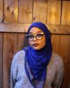 Sumayyah standing in front of a wood panel background in a blue hijab and grey sweater.