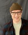 Man wearing a brown beanie, dark-framed glasses, and open flannel shirt over a light grey t-shirt stands smiling in front of a dark grey backdrop