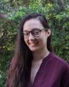 Image of Remy Sumida-Tate, a person with long dark hair wearing glasses and a maroon shirt.