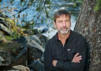 Poet Derek Sheffield, arms crossed, stands next to a tree with a river in the background.