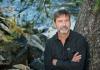 Poet Derek Sheffield, arms crossed, stands next to a tree with a river in the background.