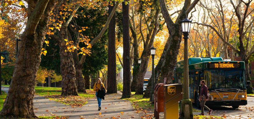 Image of campus walkway surrounded by trees with brown autumn leaves, people walking, and a bus stopped in the road